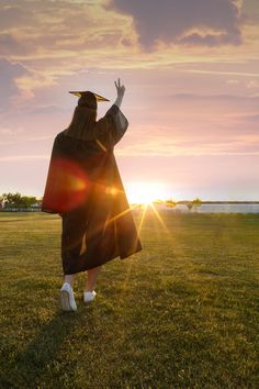 a woman in a graduation gown and hat is walking across the grass towards the sun