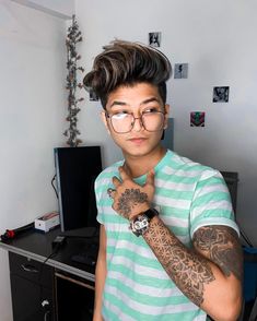 a young man with glasses and tattoos on his arm standing in front of a computer desk