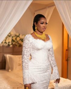 a woman standing in front of a bed wearing a white dress and yellow necklaces