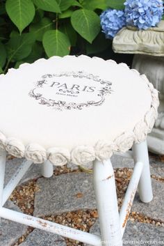 a small white stool sitting on top of a stone floor next to blue and green flowers