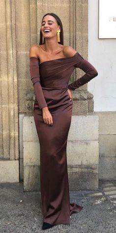 a woman in a long brown dress is laughing and standing on the sidewalk with her hands behind her head