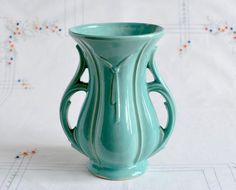 a green vase sitting on top of a white cloth covered tablecloth with flowers in the background