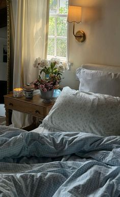 a bed with blue sheets and pillows in front of a window, next to a table with flowers on it