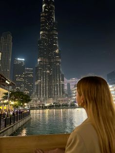 a woman looks out over the water at night in front of a cityscape