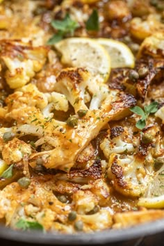 a pan filled with chicken and vegetables on top of a wooden table next to lemon wedges