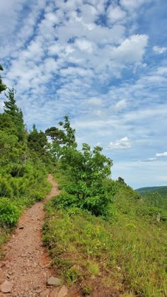 a dirt path on the side of a lush green hillside under a cloudy blue sky