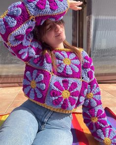 a woman sitting on the ground wearing a purple and yellow crochet sweater with flowers