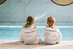 two young children sitting on the ground in front of a swimming pool wearing white robes