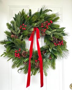 a christmas wreath with red ribbon hanging on the front door, decorated with holly and pine cones