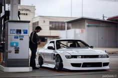 a man standing next to a white car in front of a gas station pump machine