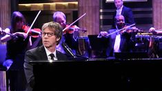 a man in a suit and tie playing the piano with other musicians behind him on stage