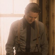 a man with suspenders and a bow tie standing in front of a window looking down