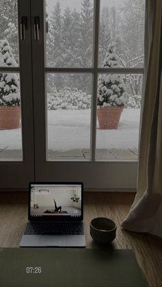 an open laptop computer sitting on top of a wooden desk next to a window covered in snow