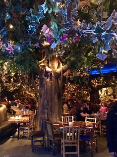 people are sitting at tables under a tree in the middle of a room with lots of lights