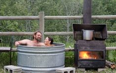 a man and woman sitting in a metal tub next to an open fire place with a wood burning stove