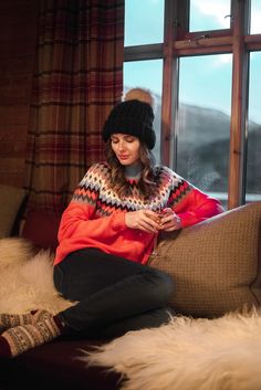 a woman sitting on a couch looking at her cell phone while wearing a knitted hat