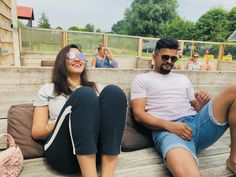 two people sitting on a wooden bench with their feet up and one person wearing sunglasses