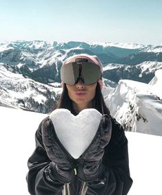 a woman wearing goggles holding a heart shaped pillow in front of her face on top of a snow covered mountain