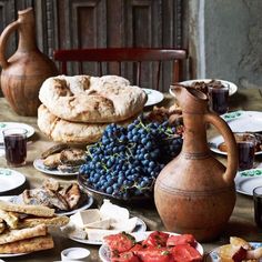 a table full of food including breads, grapes and other foods on plates next to a pitcher