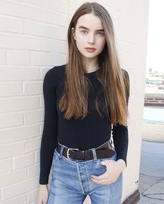 a woman standing next to a white brick wall with her hands in her pockets and looking at the camera