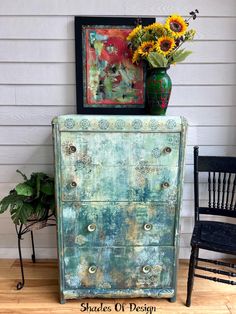 an old dresser with sunflowers and a painting on it next to a chair