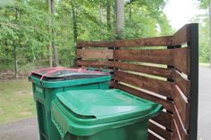 there is a green trash can next to a wooden fence and some trees in the background