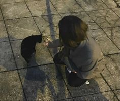 a woman kneeling down next to a black cat on top of a tiled floor and eating something off of her hand