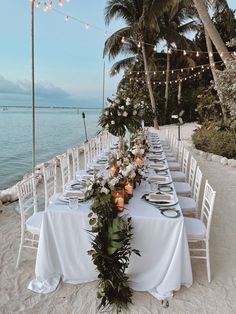 a long table set up on the beach with white linens and greenery for an elegant dinner