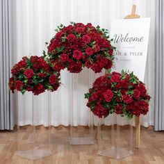 three clear vases filled with red flowers on top of a wooden floor next to a welcome sign