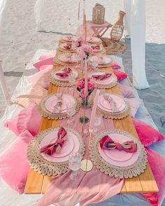 a table set up on the beach with pink and gold plates, napkins and place settings