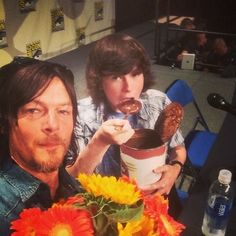 two people sitting at a table with flowers in front of them and one holding a cup