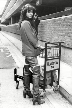 a woman standing next to a shopping cart on the side of a building with her hand in her pocket