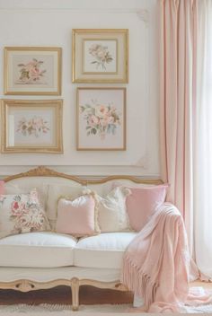 a white couch with pink and gold pillows in front of framed pictures on the wall