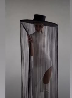 a woman in white is holding a black hat and standing behind a sheered curtain