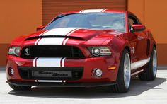 a red and white mustang car parked in front of a garage door with an orange building behind it