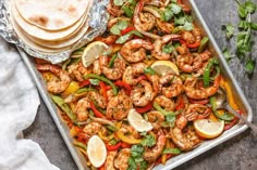 a sheet pan filled with shrimp, peppers and lemons next to tortillas