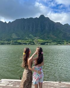 two women standing on a dock looking at the water with mountains in the background and clouds in the sky