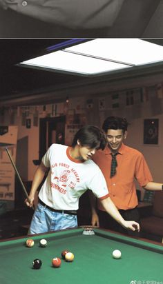 two people standing around a pool table with balls on it and one person holding a cue