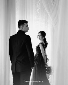 a man and woman standing next to each other in front of a window with white curtains