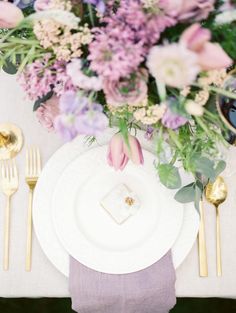 the table is set with white plates and silverware, gold cutlery, and purple flowers