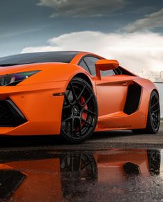 an orange sports car parked in front of a fence on a wet road with dark clouds behind it