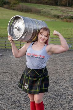 a woman in a kilt is holding a large metal barrel and posing for the camera