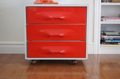 a red dresser with three drawers in front of a white wall and bookshelf