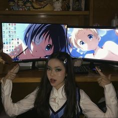 a woman with long black hair sitting in front of two computer monitors holding up her hands