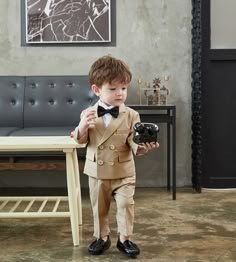 a young boy dressed in a suit and bow tie holding a black ball glove while standing next to a couch