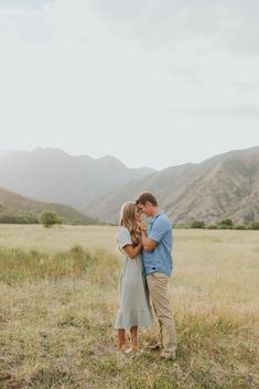 an engaged couple standing in the middle of a field with mountains in the background at sunset