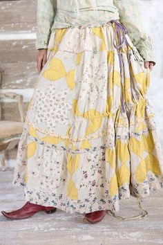 a woman wearing a yellow and white dress with flowers on the skirt is standing in front of a wooden wall