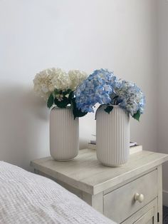 two white vases with blue and white flowers in them on a table next to a bed