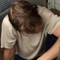 a man with long hair sitting on the floor in front of a sink and looking down at his cell phone