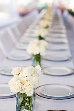 a long table with white flowers in a vase and plates on the side, along with place settings